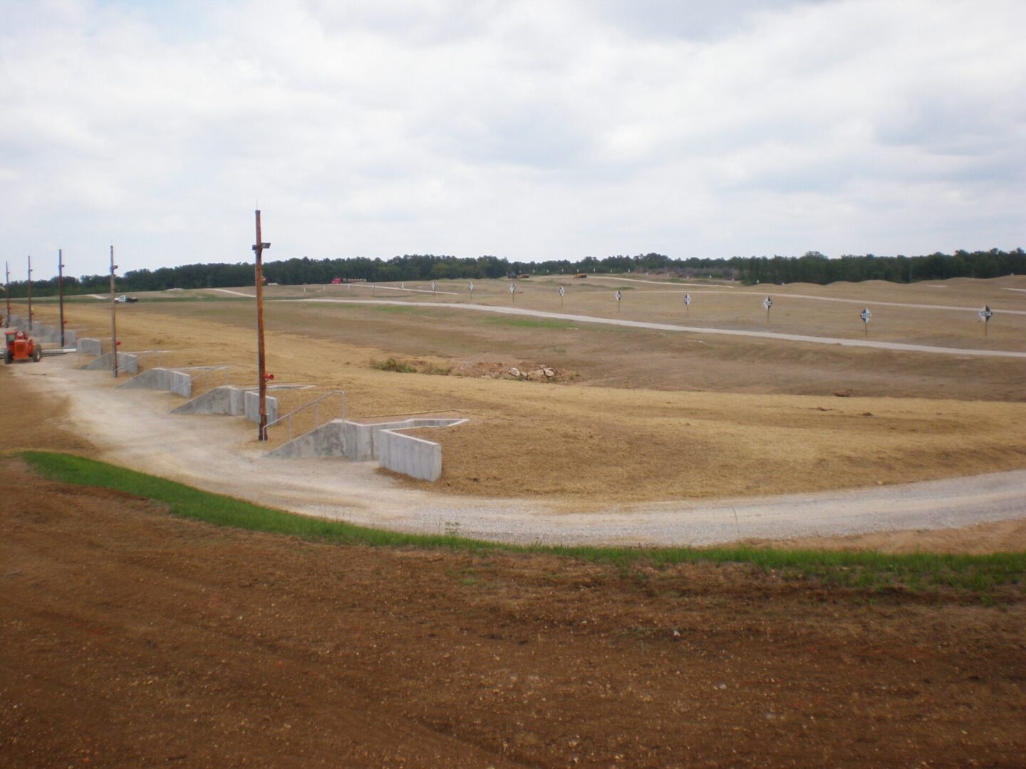 A view of ongoing construction at a large, open site with a curved dirt road, concrete barriers, and utility poles under a cloudy sky.