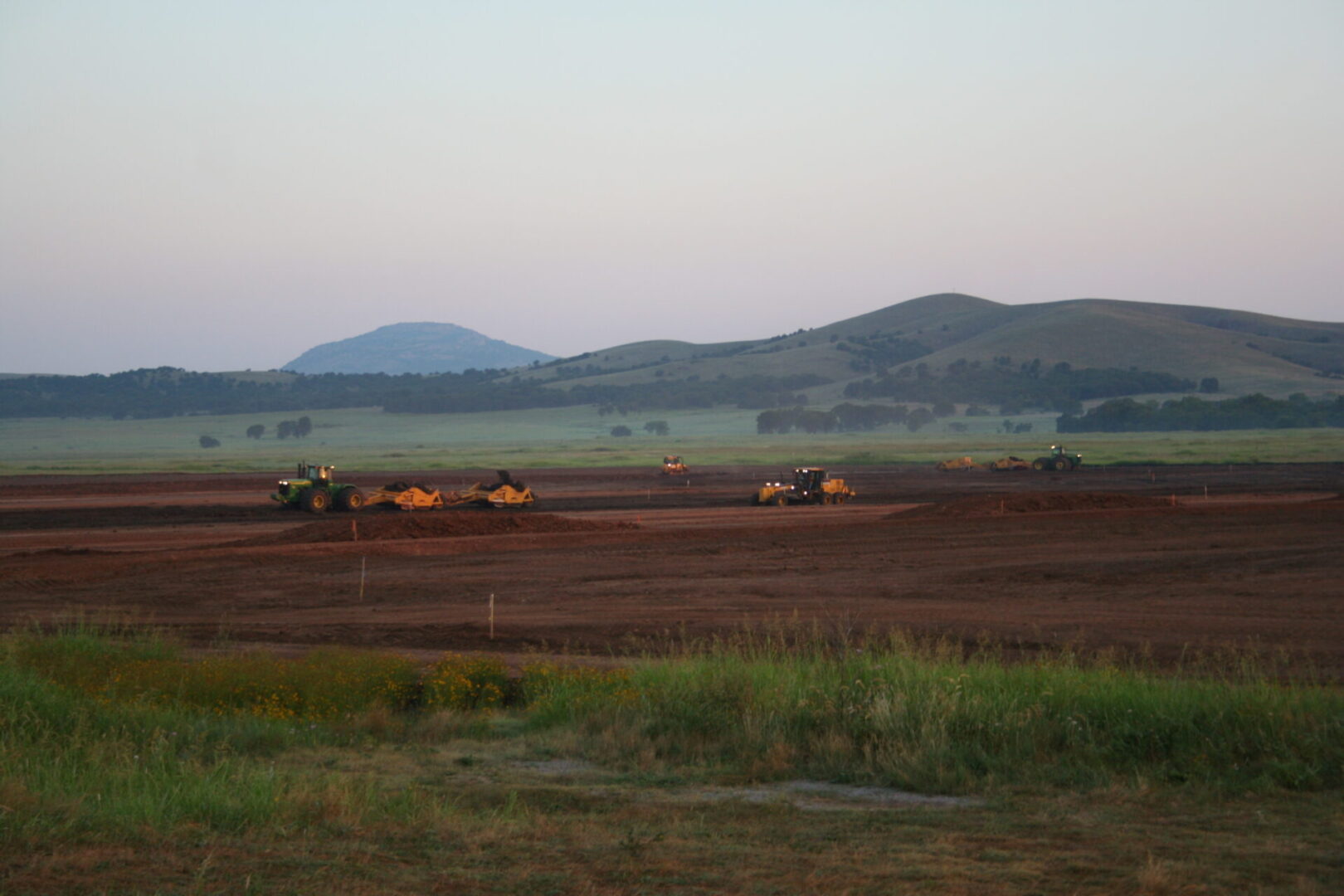 Tractors working on a construction site at dusk with hills in the background.